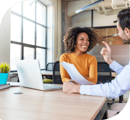 A sitting woman in front of a laptop smiles at a man as they talk about small business banking